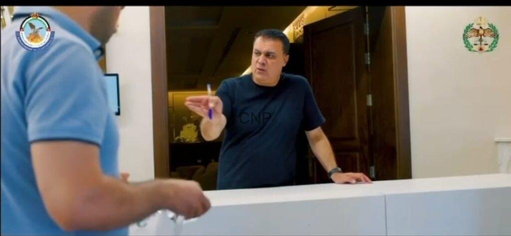 bb1 - Center News Network Man at a reception counter points a pen toward a visitor in a blue shirt, logos visible on the walls behind him.
