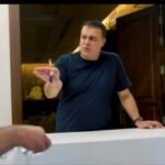 Man at a reception counter points a pen toward a visitor in a blue shirt, logos visible on the walls behind him.