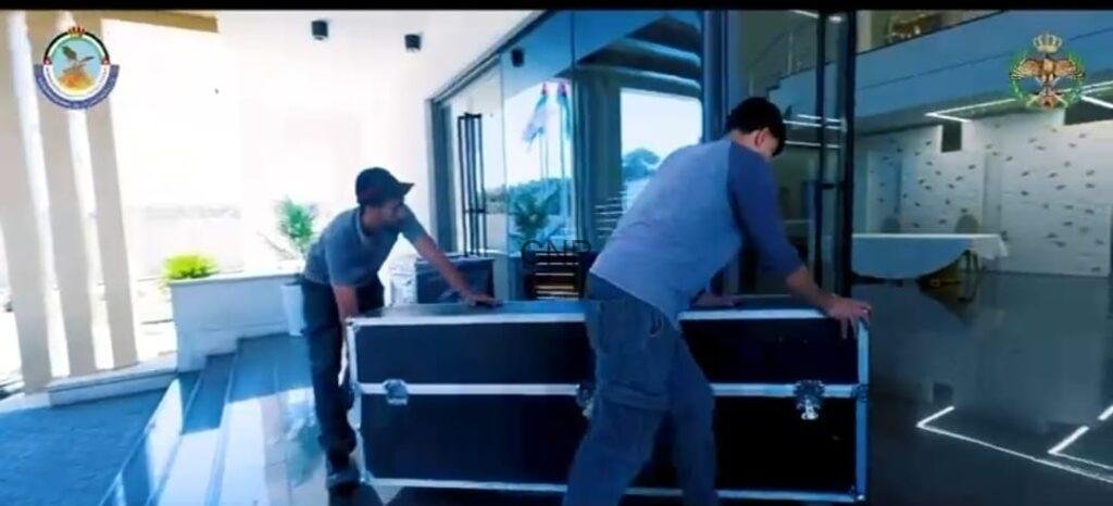 Two men move a large black flight case across a tiled outdoor area in front of a glass-building entrance, preparing for transport.