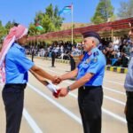 Two uniformed officers shake hands during a formal ceremony on a parade ground, with a crowd watching under a striped canopy and flags in the background.