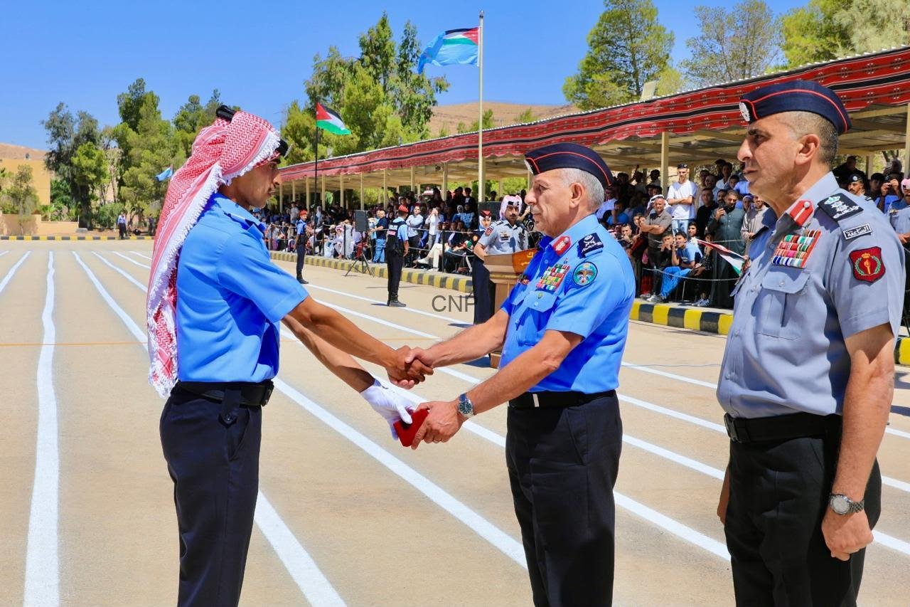 Two uniformed officers shake hands during a formal ceremony on a parade ground, with a crowd watching under a striped canopy and flags in the background.