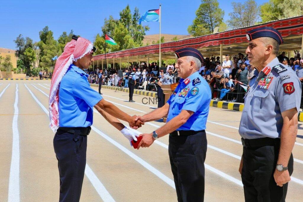 haf1 - Center News Network Man in a blue uniform with a red-and-white keffiyeh shakes hands with a fellow officer on a parade ground as spectators watch from a shaded stand behind barriers.