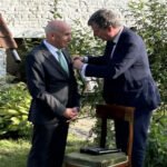 Two men in suits outdoors; one pins a boutonniere on the other's lapel at a garden ceremony.