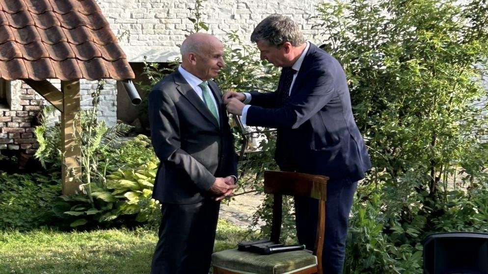 Two men in suits outdoors; one pins a boutonniere on the other's lapel at a garden ceremony.