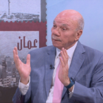 Bald man in a light blue suit and pink tie speaks on a TV set, gesturing with both hands while wearing a lapel mic.