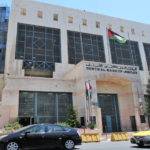 Facade of the Central Bank of Jordan building with a Jordanian flag above the entrance and cars on the street in front.