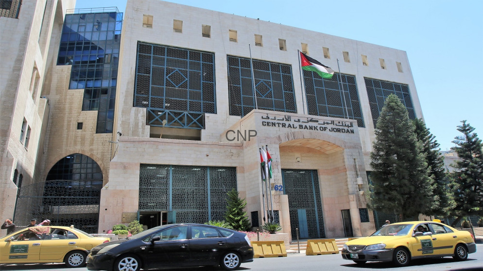 Facade of the Central Bank of Jordan building with a Jordanian flag above the entrance and cars on the street in front.