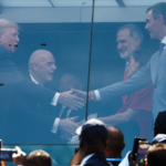 Business leaders shake hands through a glass partition at a public event, with a cheering crowd taking photos in the foreground.