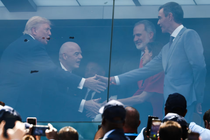 Business leaders shake hands through a glass partition at a public event, with a cheering crowd taking photos in the foreground.