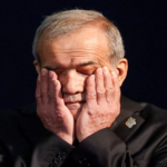Older man with gray hair holding his face in his hands on a dark stage, eyes closed in thought or fatigue.