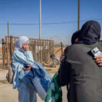 Two women wearing headscarves hug and take a selfie near a fenced border area in a desert setting.