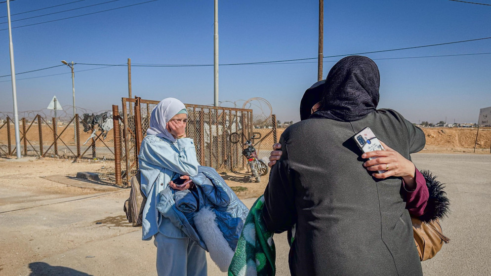 image - Center News Network Two women wearing headscarves hug and take a selfie near a fenced border area in a desert setting.
