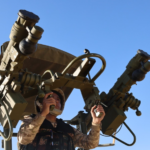 Military service member in desert camouflage operating a large dual-barrel mounted weapons system outdoors under a blue sky