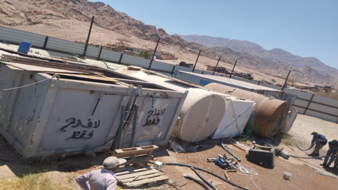 Desert industrial yard with rusted cylindrical tanks and metal containers, ladders, and scattered hoses, with two workers in the foreground and mountains in the distance.
