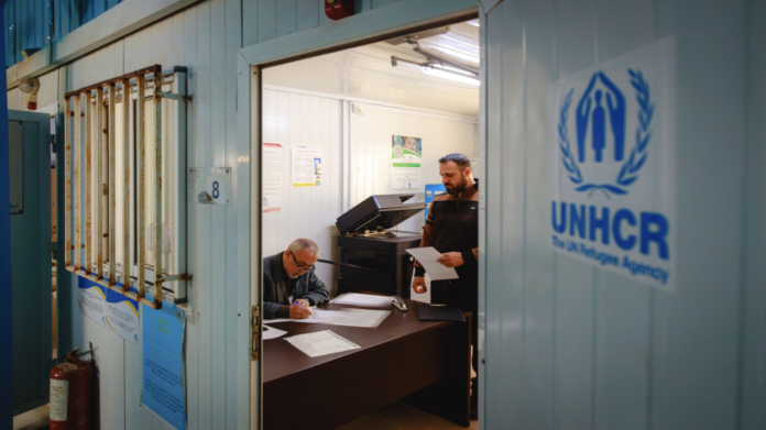 Two men in a small office signing and reviewing documents, with the UNHCR logo on the blue door outside the room.