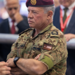 Older man in camouflage uniform with a maroon beret and a gold insignia, arms crossed, in a crowd scene.