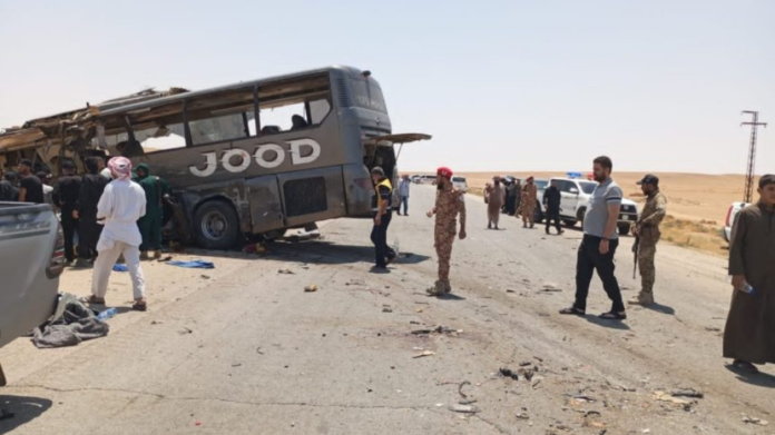 Bus overturned on a desert highway after a crash; bystanders and security personnel assess the scene.