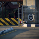 Entrance to a secured facility with a tall metal gate and a yellow‑and‑black striped barrier; a guard stands by a car in the background.