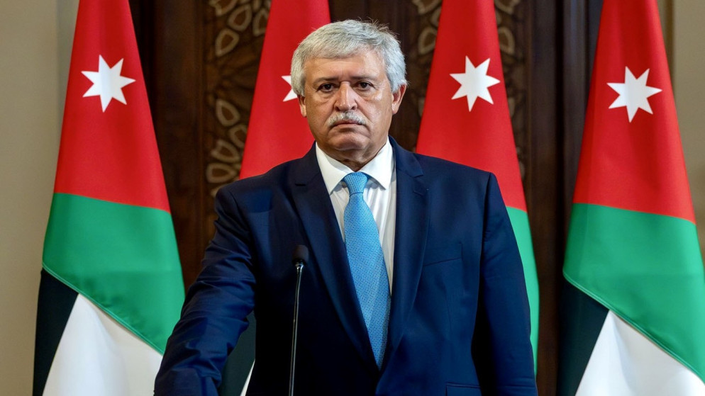 Middle-aged man in a navy suit and light blue tie standing at a podium, with Jordanian flags in the background.