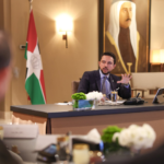Jordanian official speaks at a formal meeting table with the flag and portrait in the background, attendees blurred in foreground