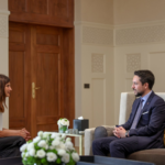 Two people sit facing each other in a formal office: a woman in a white blouse and a man in a navy suit, with a Jordanian flag in the background.