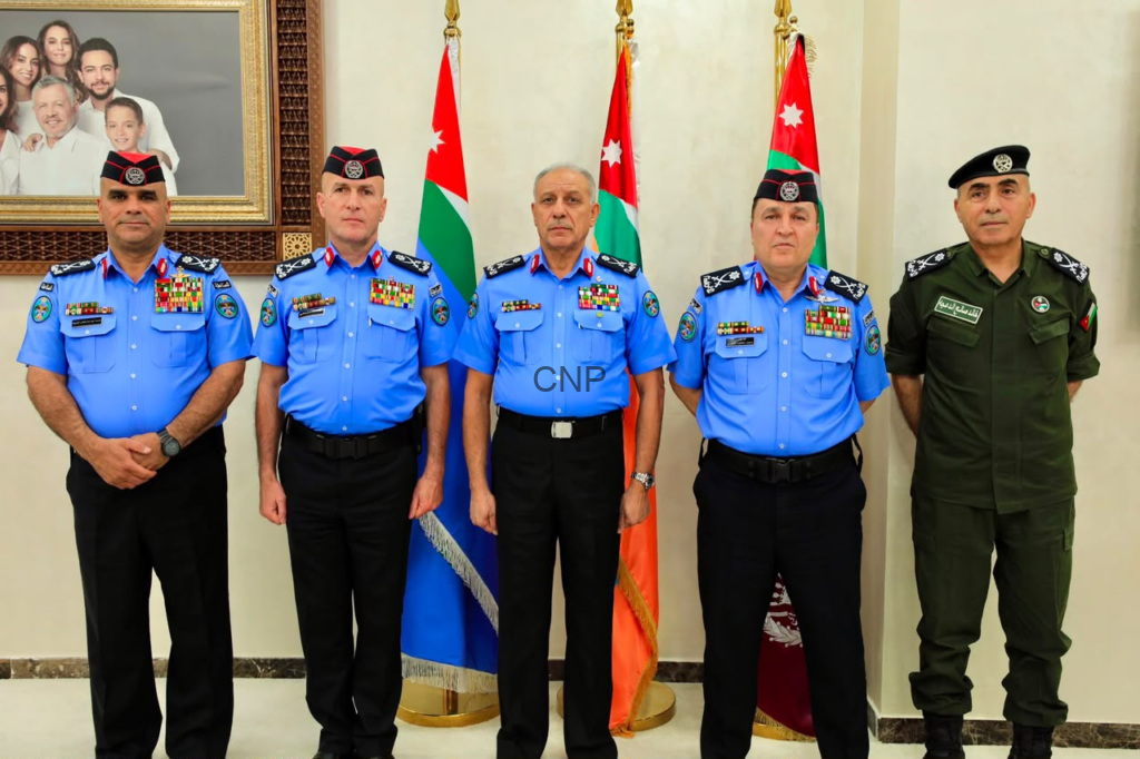 Five uniformed men stand in a row indoors, four in light-blue dress uniforms and one in green, with Jordanian flags behind them and a framed family portrait on the wall to the left.