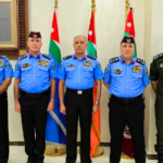 Five uniformed men stand in a row indoors, four in light-blue dress uniforms and one in green, with Jordanian flags behind them and a framed family portrait on the wall to the left.