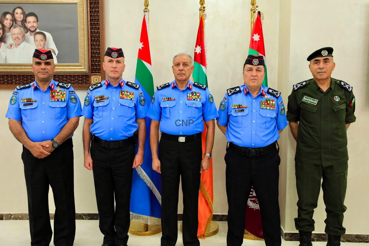 Five uniformed men stand in a row indoors, four in light-blue dress uniforms and one in green, with Jordanian flags behind them and a framed family portrait on the wall to the left.