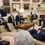 Group of world leaders seated in a gilded, portrait-filled parlor, discussing around a central coffee table.