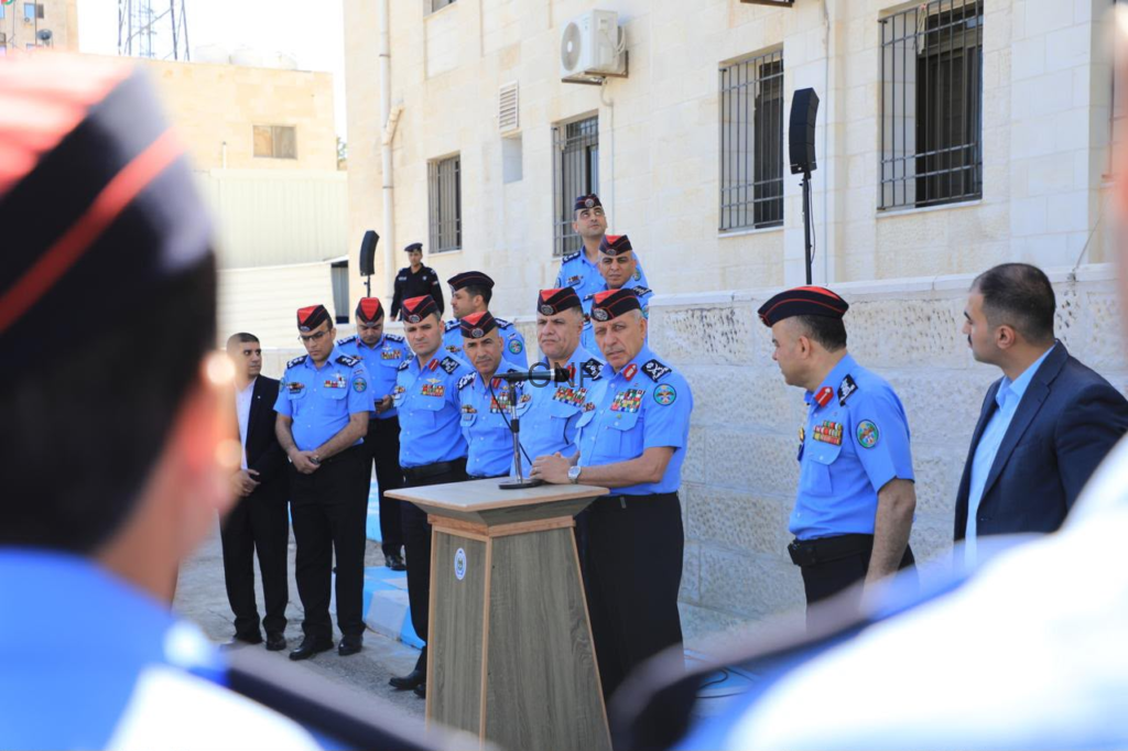 image - Center News Network Police officers in blue uniforms stand at a wooden podium giving a speech outdoors, with a row of officials and onlookers behind them in a courtyard building setting