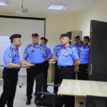 Group of police officers in blue uniforms and red berets standing in a classroom, one officer speaking as others listen.