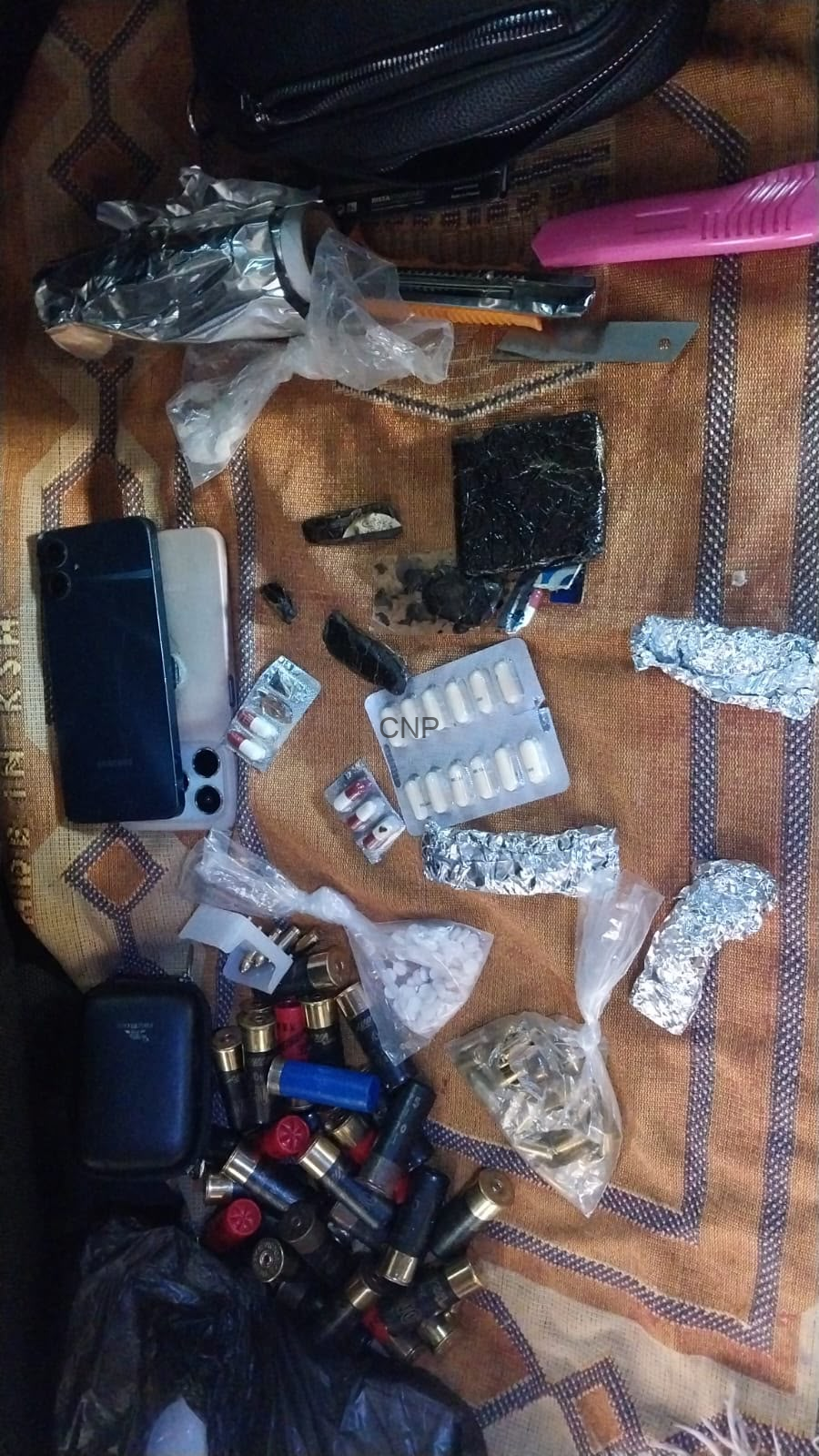 Assorted drug paraphernalia on a patterned cloth: blister packs, pills, foil packets, plastic bags, and a phone nearby.