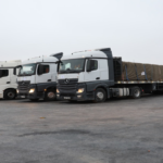 Row of white semi-trucks parked in a lot, connected to long trailers covered with tarps on a gray day