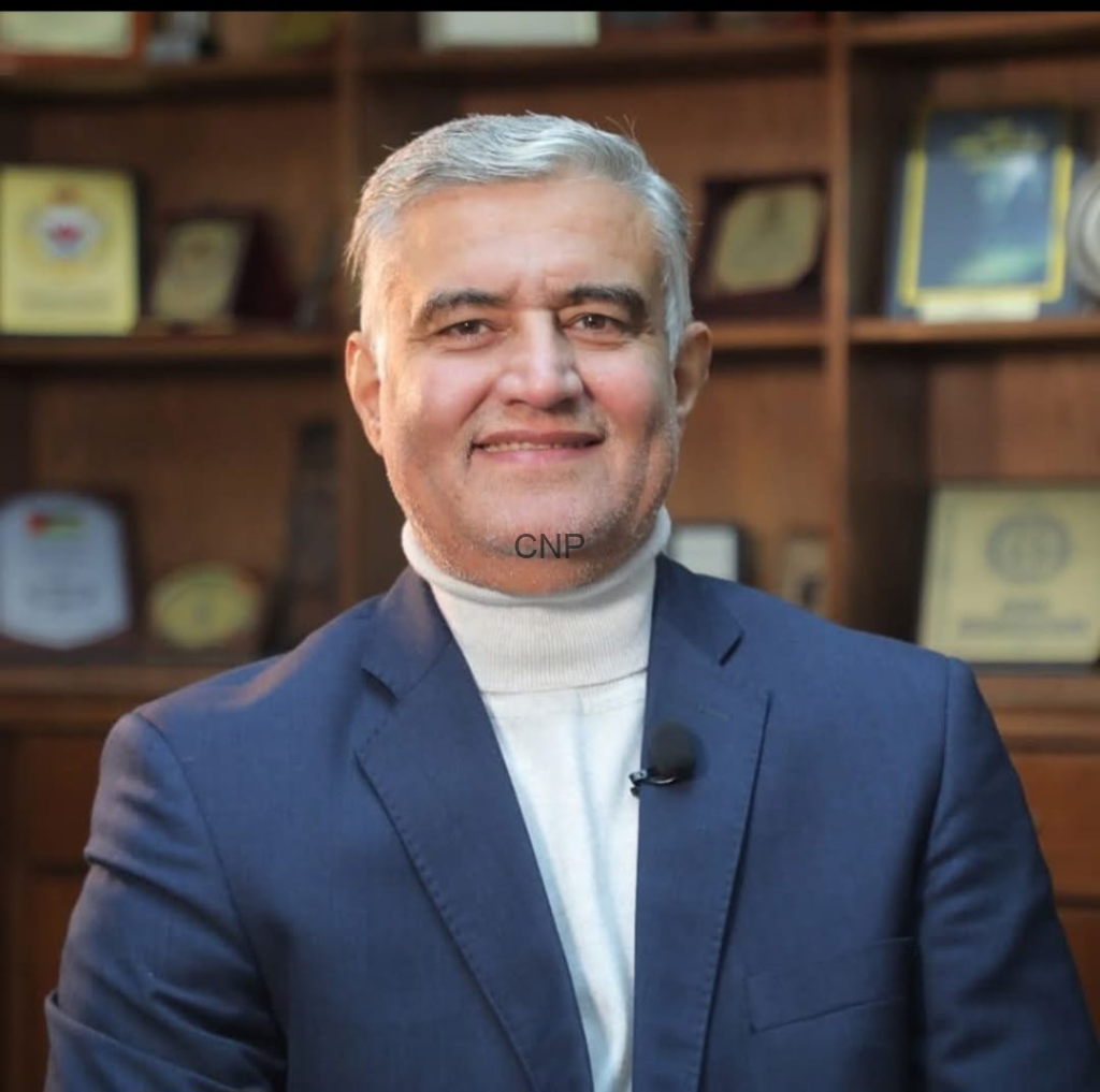 image - Center News Network Smiling middle-aged man in a navy suit with a cream turtleneck, mic clipped to his lapel, in an office with wooden shelves and awards in the background.