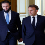 Two men in dark suits walk out of an ornate doorway, one wearing a blue striped tie, the other with a black tie, looking ahead.