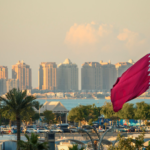 Qatar flag waving beside a waterfront skyline with tall buildings and palm trees in the foreground.