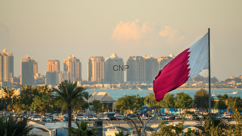 image - Center News Network Qatar flag waving beside a waterfront skyline with tall buildings and palm trees in the foreground.