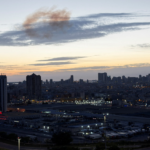 Dusk city skyline with tall buildings and an industrial foreground under a orange-blue sunset.