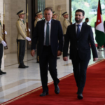 Two men in suits walk on a red carpet during a formal welcome, flanked by guards with rifles; a flag is visible in the background.