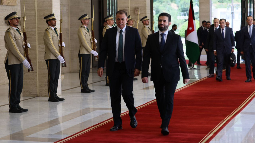 Two men in suits walk on a red carpet during a formal welcome, flanked by guards with rifles; a flag is visible in the background.
