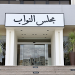Entrance to a government building with a prominent Arabic sign reading مجلس النواب above glass doors, flanked by tall planters