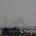 Urban skyline with a distant plume of smoke rising over the horizon; foreground shows a multi-story building with an antenna.