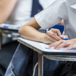 Students in a classroom taking notes at desks, writing on papers with blue pens and notebooks visible in the foreground.