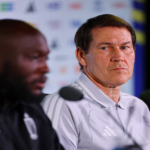 Two men at a press conference with microphones; focus on the man in a light gray Adidas polo looking serious, background logos blurred.