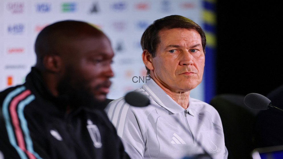 image - Center News Network Two men at a press conference with microphones; focus on the man in a light gray Adidas polo looking serious, background logos blurred.