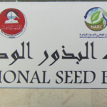 Signboard for National Seed Bank with Arabic text and two circular logos above the English title on a pale wall.