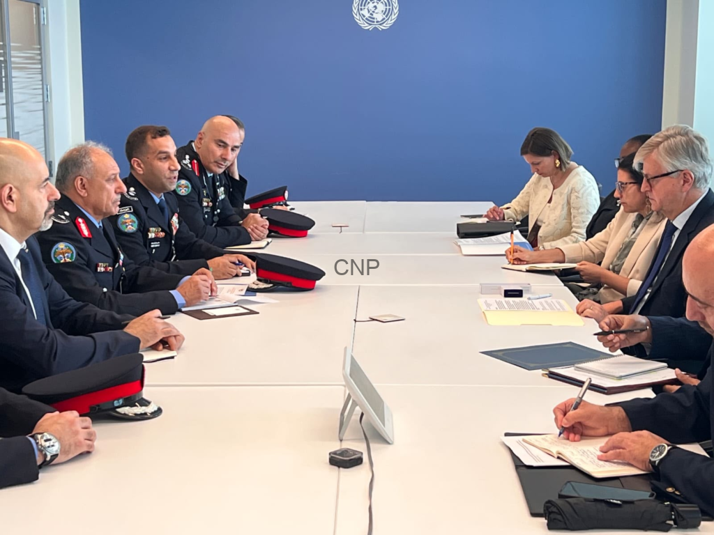 Military officers and civilian diplomats sit around a long conference table in a United Nations meeting room, blue wall with UN emblem behind them, taking notes and discussing.