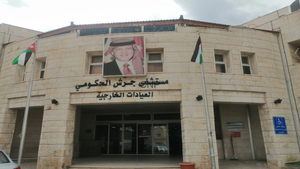 image - Center News Network Exterior of Jerash Government Hospital with a portrait of the Jordanian king and Jordanian flags at the entrance; Arabic sign naming outpatient clinics.