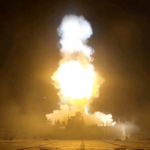 Rocket engine test firing on a ground stand at night with a large bright exhaust plume and surrounding equipment.