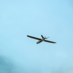 Small propeller airplane seen from below, flying against a pale blue sky.